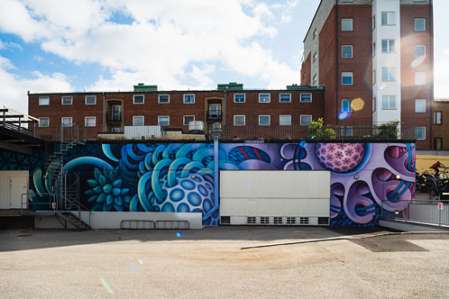 Muralmålning med abstrakta geometriska former, mönster och illusioner av rumslighet i varma jordfärger. Konstverket täcker en lång fasad med garageportar i kvarteret Gjutaren i Eslöv.