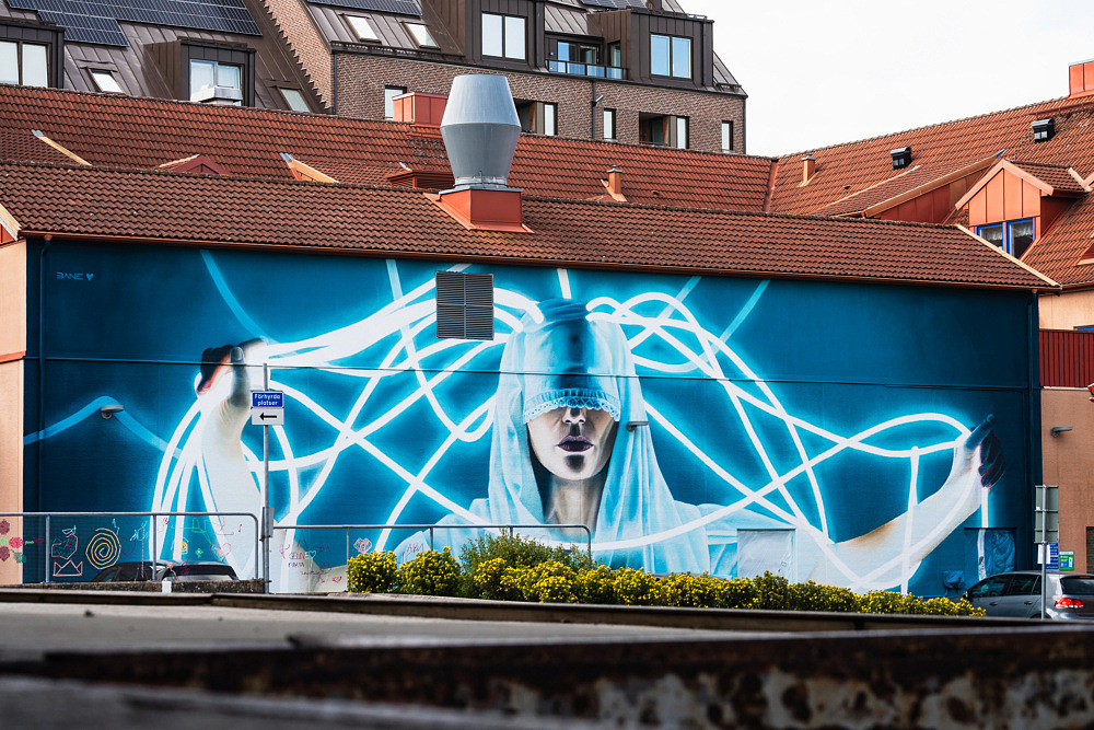 Fotorealistisk muralm&aring;lning av en ung kvinna som blickar mot betraktaren. Portr&auml;ttet t&auml;cker en h&ouml;g husfasad och &auml;r utf&ouml;rt i mjuka f&auml;rgtoner med detaljerade skuggor och ljuseffekter.