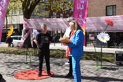 Tre personer st&aring;r p&aring; en scen p&aring; Stora torg i Esl&ouml;v under en prisutdelning i str&aring;lande solsken. En kvinna i bl&aring; kostym h&aring;ller tal i mikrofon medan tv&aring; personer bredvid h&aring;ller ett diplom och en stor prischeck.