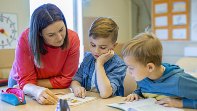 två förskolebarn, pojkar, sitter vid ett bord och talar med en kvinnlig förskolelärare