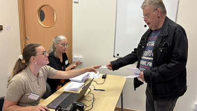 Man lämnar över dokument till två kvinnor som sitter vid ett bord med datorer framför sig.