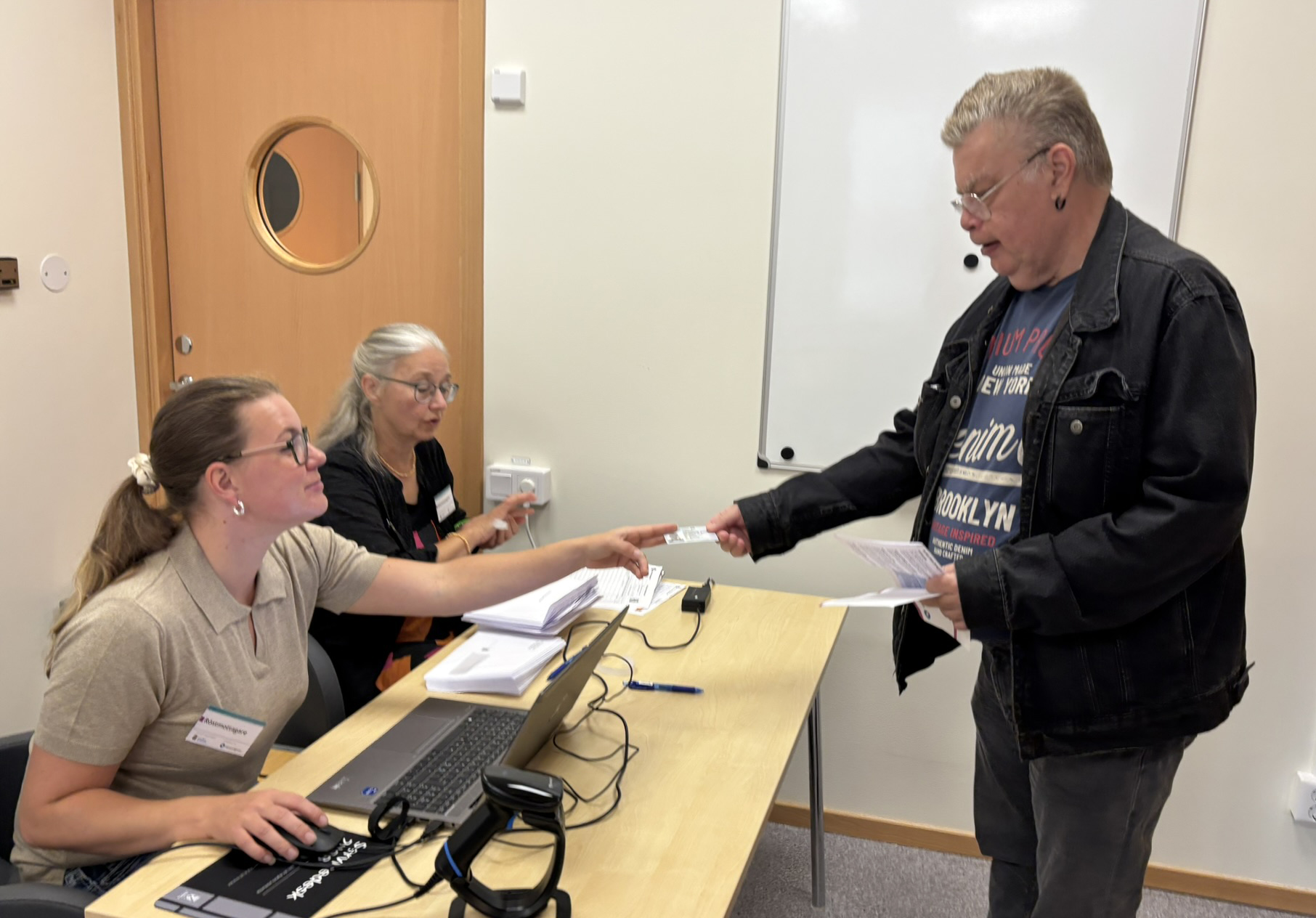Man l&auml;mnar &ouml;ver dokument till tv&aring; kvinnor som sitter vid ett bord med datorer  framf&ouml;r sig.