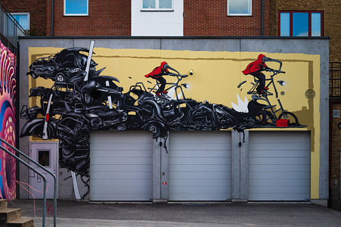 Muralmålning med svarta bilar i olika former på ljusgul bakgrund. Två personer i röda kappor cyklar genom motivet som sträcker sig längs en lång fasad med garageportar.