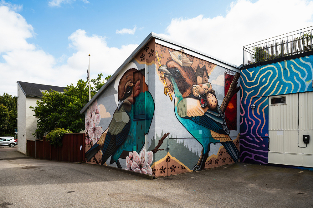 F&auml;rgstark muralm&aring;lning med tv&aring; stiliserade f&aring;glar som m&ouml;ts p&aring; ett hustorn. F&aring;glarna b&auml;r kl&auml;der och st&aring;r i ett poetiskt stadsliknande landskap m&aring;lat &ouml;ver tv&aring; sammanh&auml;ngande fasader.