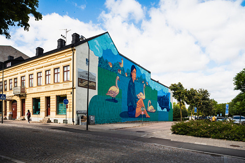 Färgstark väggmålning som föreställer en kvinna i ett drömlikt landskap med järnvägsspår. Målningen täcker en hel husfasad i centrala Eslöv.