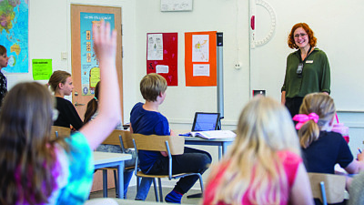 En kvinnlig lärare står framför en klass med barn som sträcker upp händerna