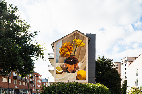 Muralmålning med en vas fylld av blommor och citroner i varma jordnära färger. Konstverket täcker en hög husgavel i centrala Eslöv.