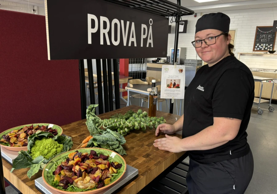 Svartklädd leende kvinnlig kock vid bord med färgglada grönsaker.