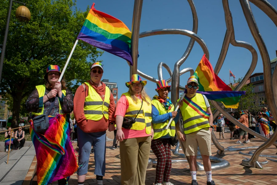 Fem personer st&aring;r tillsammans p&aring; ett torg och h&aring;ller i tv&aring; flaggor med regnb&aring;gsf&auml;rger.