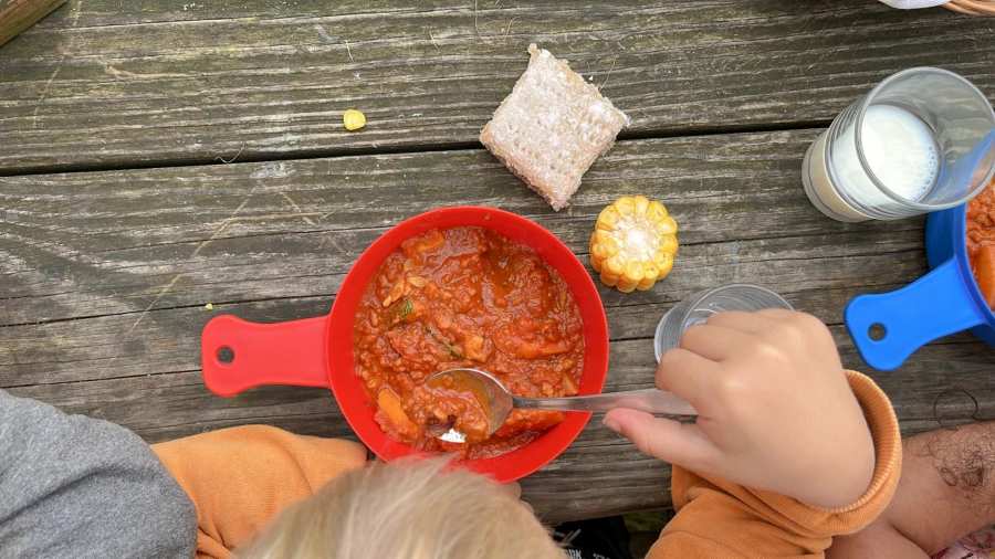 Bilden är tagen ovanifrån och visar en röd platsskål som innehåller mat. Ett barns hand håller i en matsked. Bredvid skålen, på matbordet finns en majskolv, en brödmacka och ett glas mjölk.