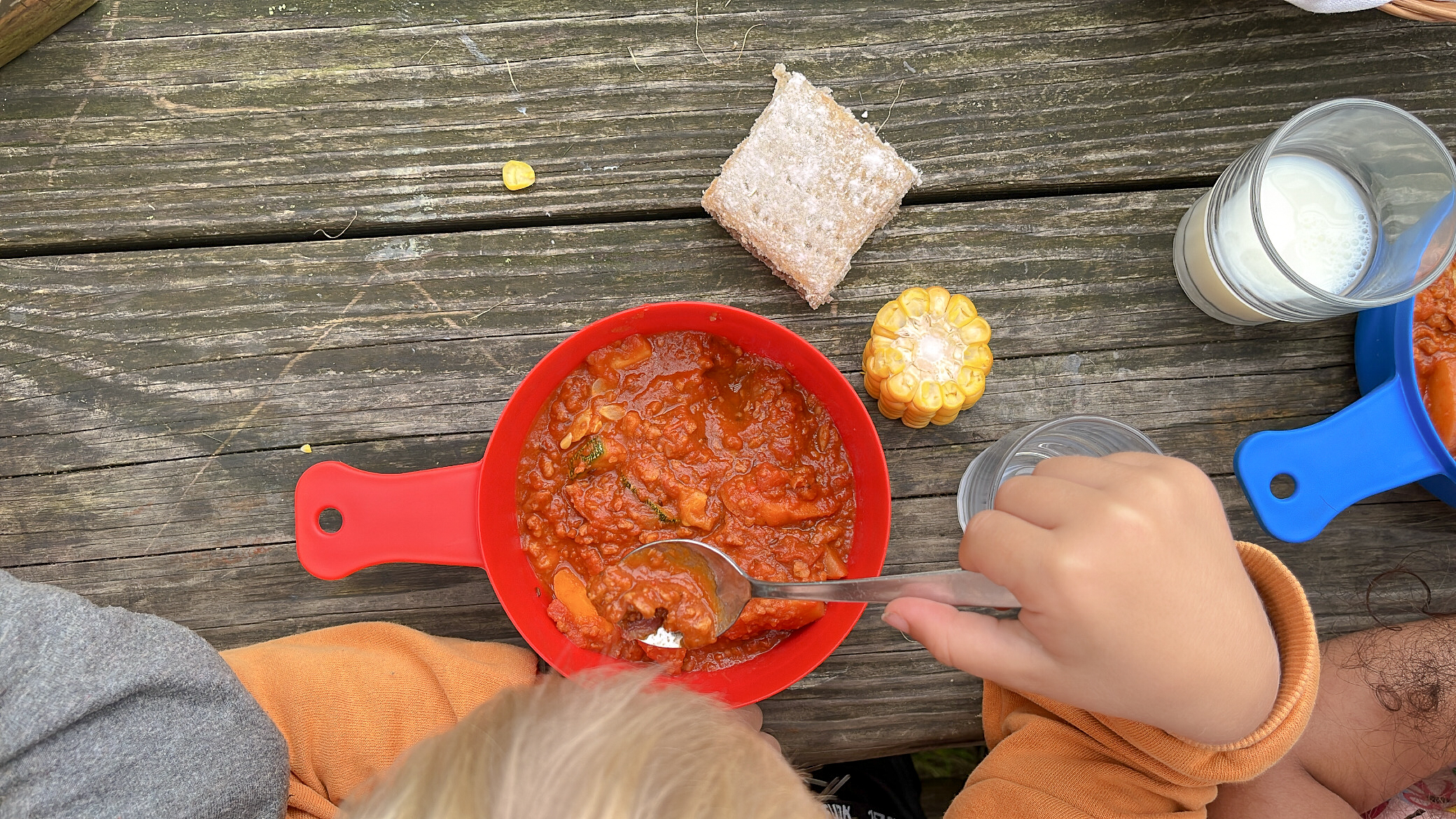 Bilden är tagen ovanifrån och visar en röd platsskål som innehåller mat. Ett barns hand håller i en matsked. Bredvid skålen, på matbordet finns en majskolv, en brödmacka och ett glas mjölk.