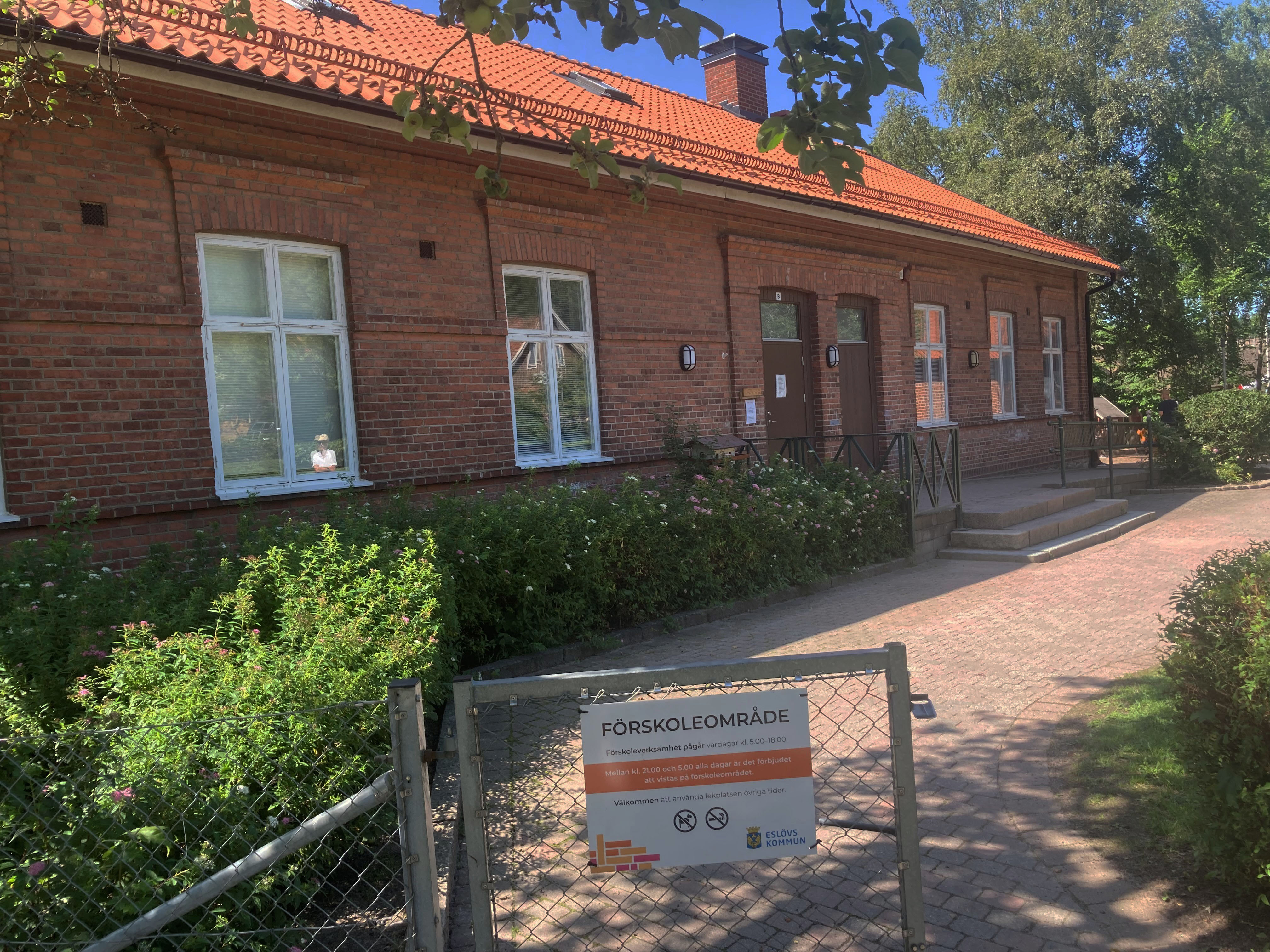 Förskola i ett plan byggt i rött tegel och med rött tegeltak. Buskar framför, grusgång oc halvöppen grind med skylt.