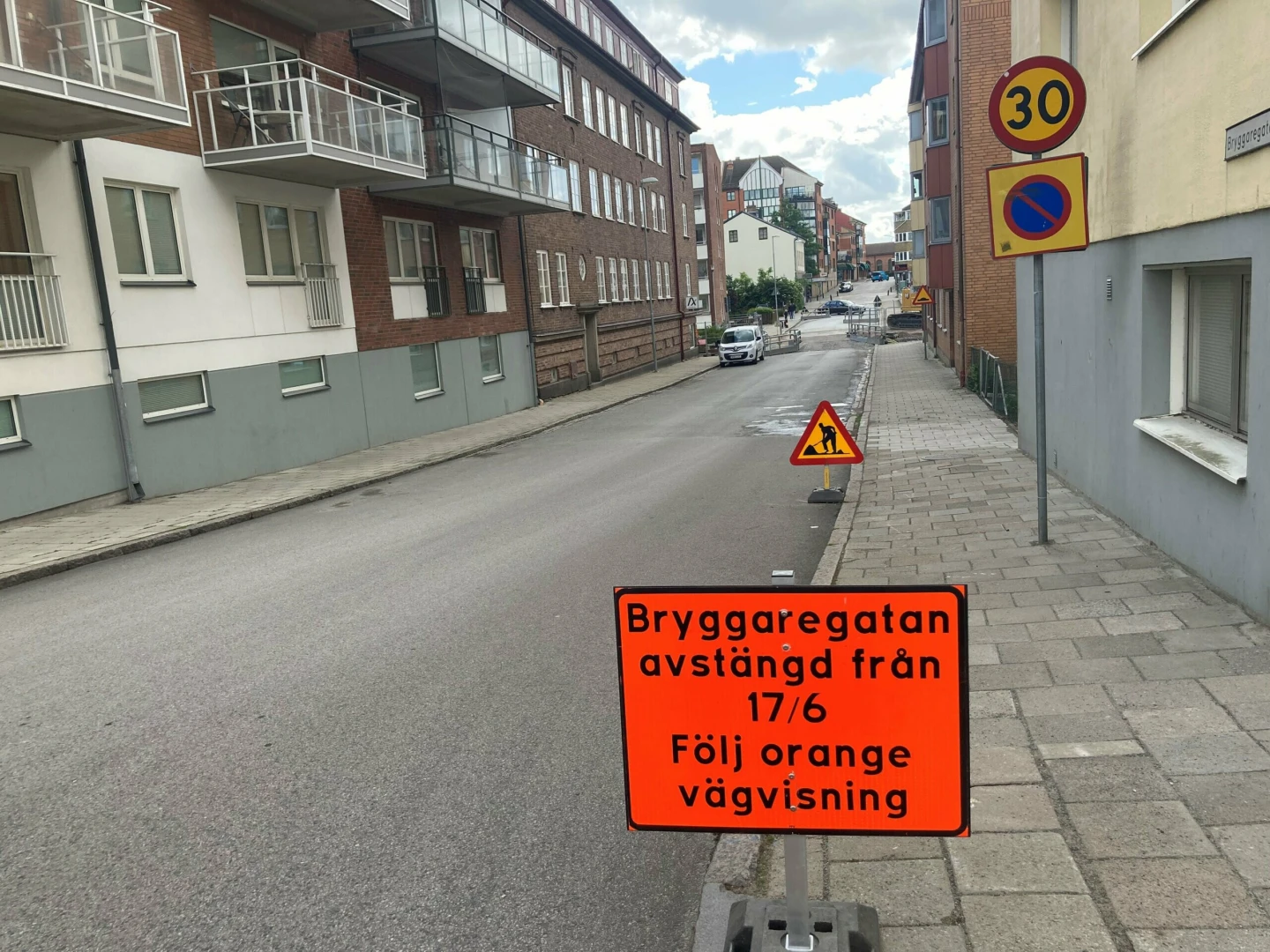 Röd skylt med text att gatan stängs av från 17/6. Tom gata och tegelhus bakom skylten.