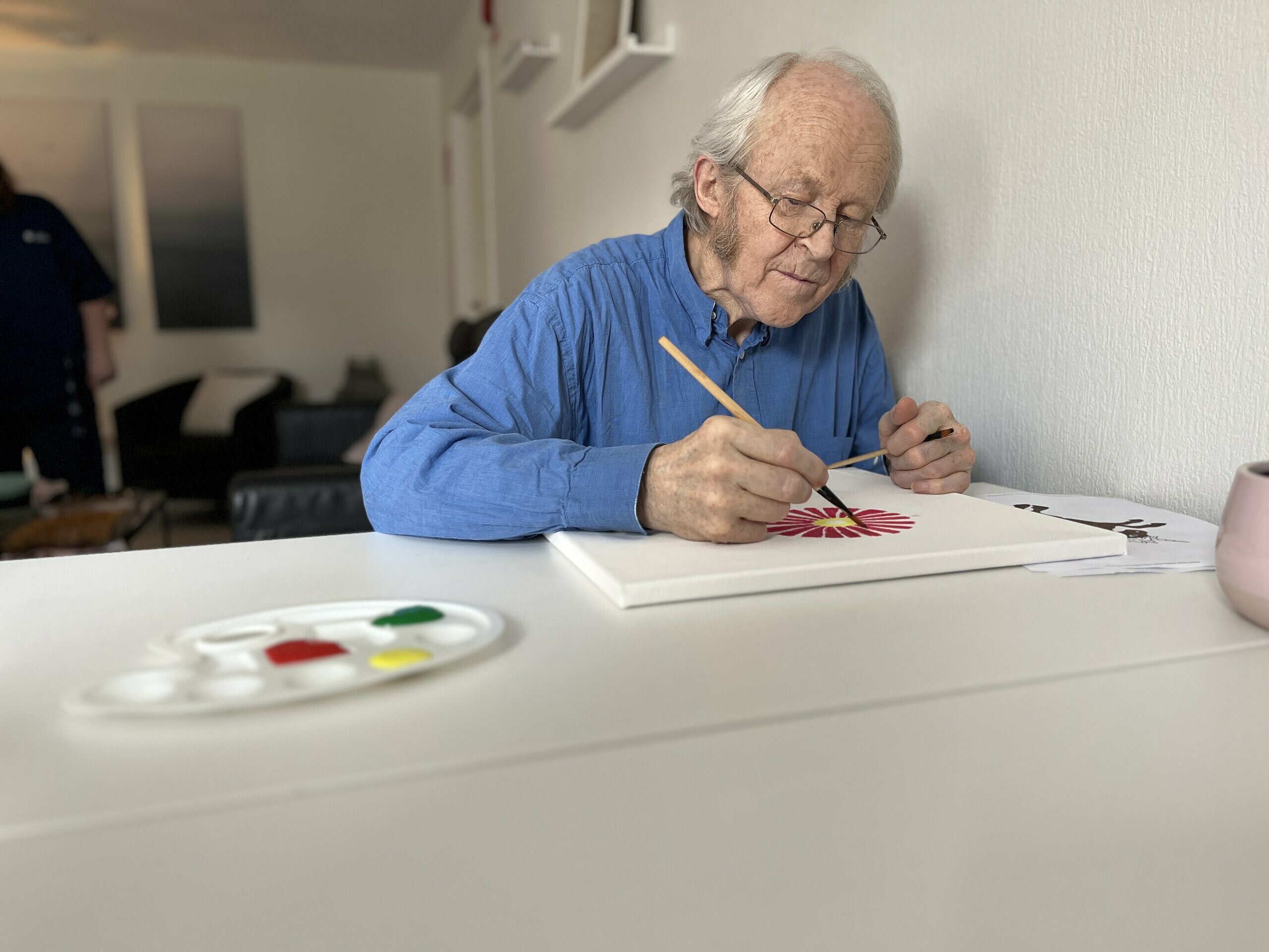 En man sitter vid matbordet och målar en tavla. Framför honom ligger en palett med tre färger: rött, gult och grönt. Han verkar måla en blomma.