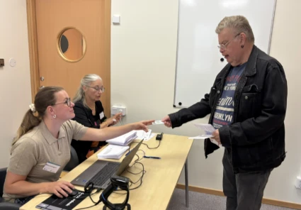 Man lämnar över dokument till två kvinnor som sitter vid ett bord med datorer framför sig.