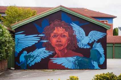 Färgstark muralmålning med ett stort porträtt målat över en hög husfasad. Konstverket använder starka färger och mjuka övergångar som skapar ett uttrycksfullt och levande motiv.