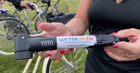 Händer håller i stafettpinnen – en cykelpump. Text på cykelpumpen: Luften är fri Friluftslivets år 2021 #skånestafetten n - en cykelpump. Text på pumpen