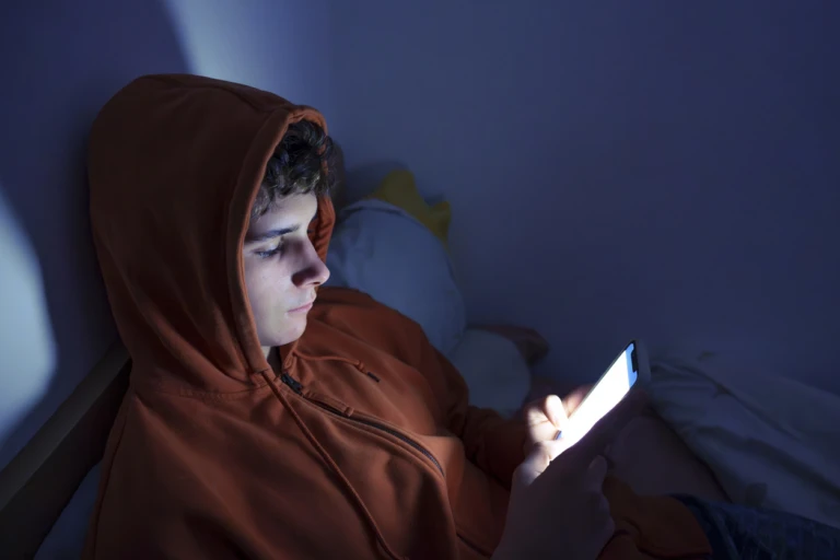 En ung kille ligger i sängen och håller på med sin mobil