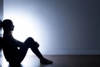Ensam person som sitter lutade mot vägg med armarna runt benen.