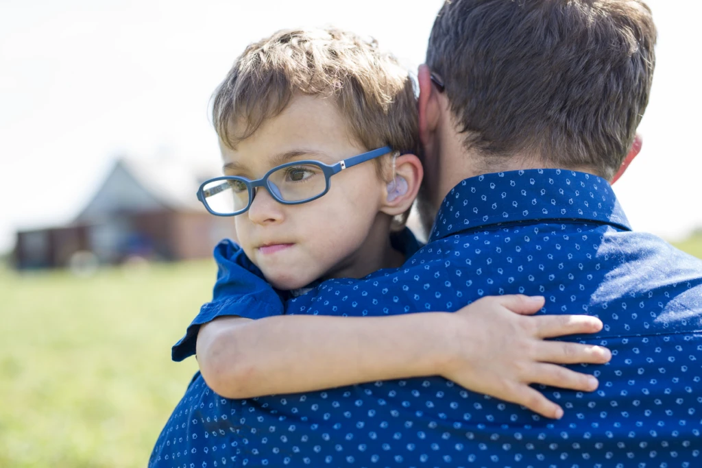 Liten pojke håller om sin pappa runt halsen. Pojken har glasögon och hörapparat