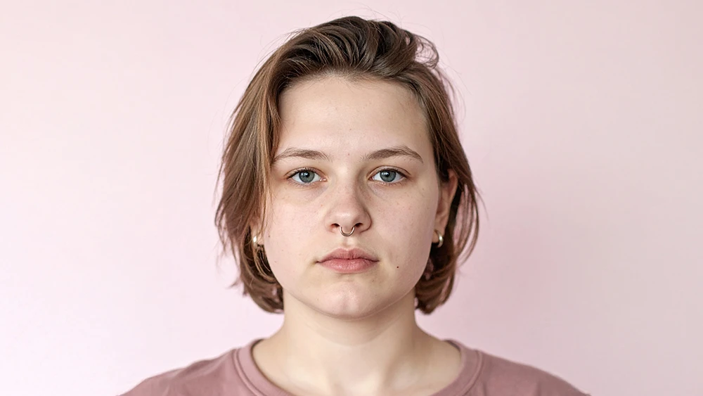 En person står framför en ljusrosa bakgrund. Personen har kort brunt hår och bär en rosa tröja.