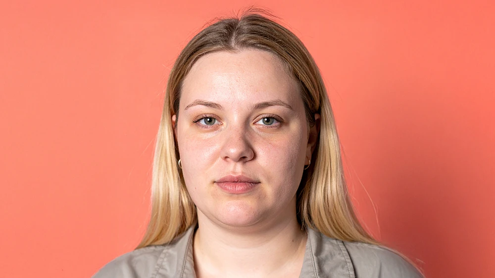 En ung kvinna porträtterad rakt framifrån mot en korallfärgad bakgrund. Hon har långt blont hår och bär en beige skjorta. Ansiktet är neutralt utan leende.