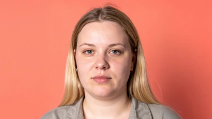 En ung kvinna porträtterad rakt framifrån mot en korallfärgad bakgrund. Hon har långt blont hår och bär en beige skjorta. Ansiktet är neutralt utan leende.