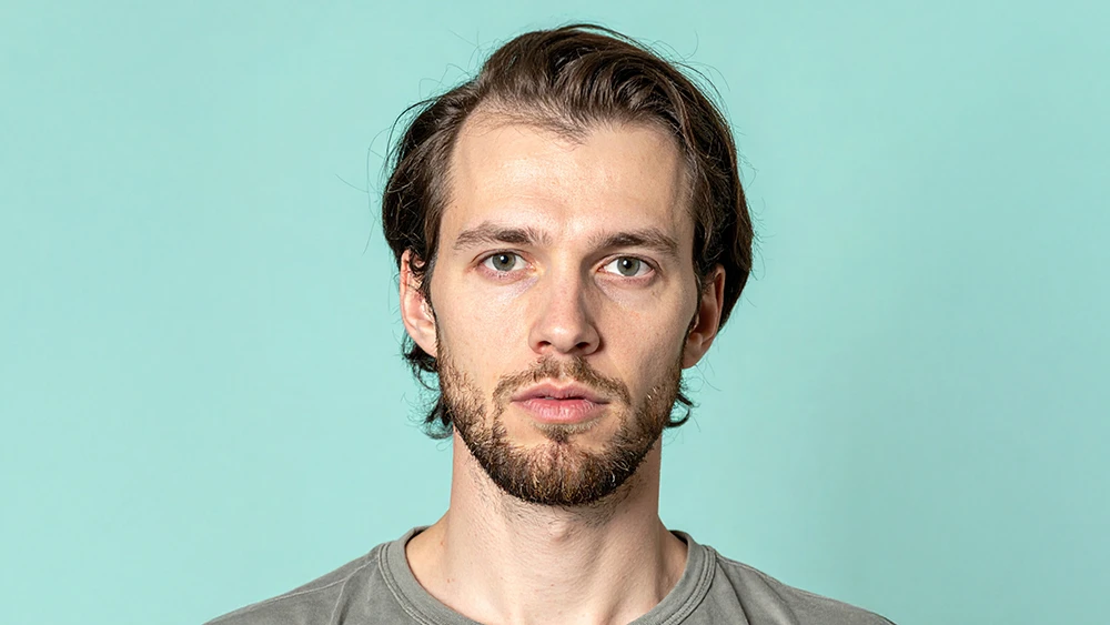 En person står framför en ljus turkos bakgrund. Personen har brunt hår och bär en grå t-shirt.