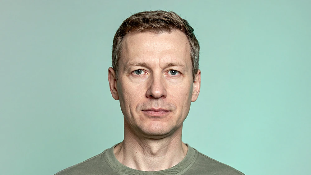 En man i femtioårsåldern porträtterad rakt framifrån mot en ljus mintgrön bakgrund. Han har kort brunt hår och bär en olivgrön t-shirt. Ansiktet är neutralt utan leende.