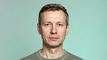 En man i femtioårsåldern porträtterad rakt framifrån mot en ljus mintgrön bakgrund. Han har kort brunt hår och bär en olivgrön t-shirt. Ansiktet är neutralt utan leende.