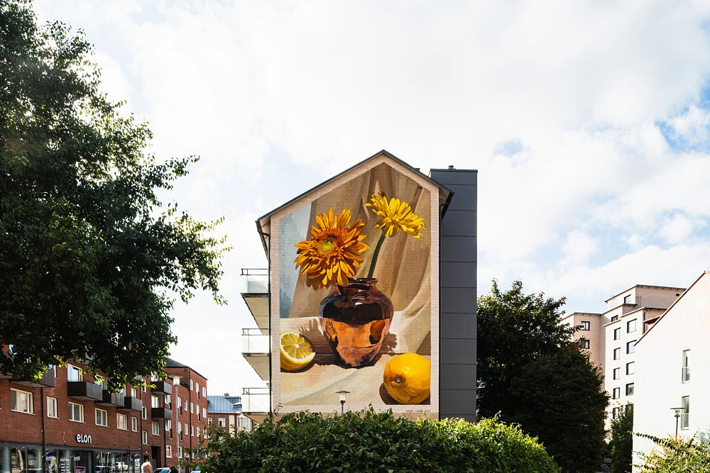 Muralmålning med en vas fylld av blommor och citroner i varma jordnära färger. Konstverket täcker en hög husgavel i centrala Eslöv.