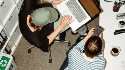 Två personer arbetar med en bärbar dator och en mobiltelefon vid ett skrivbord.