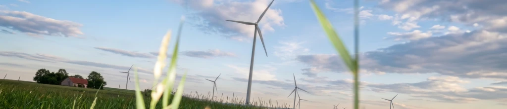 Vindkraftverk i ett öppet jordbrukslandskap med högt gräs och en gård i bakgrunden.