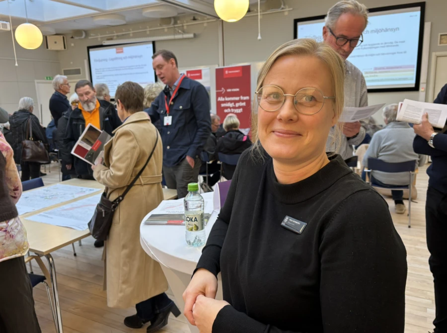 Leende ljush&aring;rig kvinna vid ett st&aring;bord. I bakgrunden flera m&auml;nniskor.