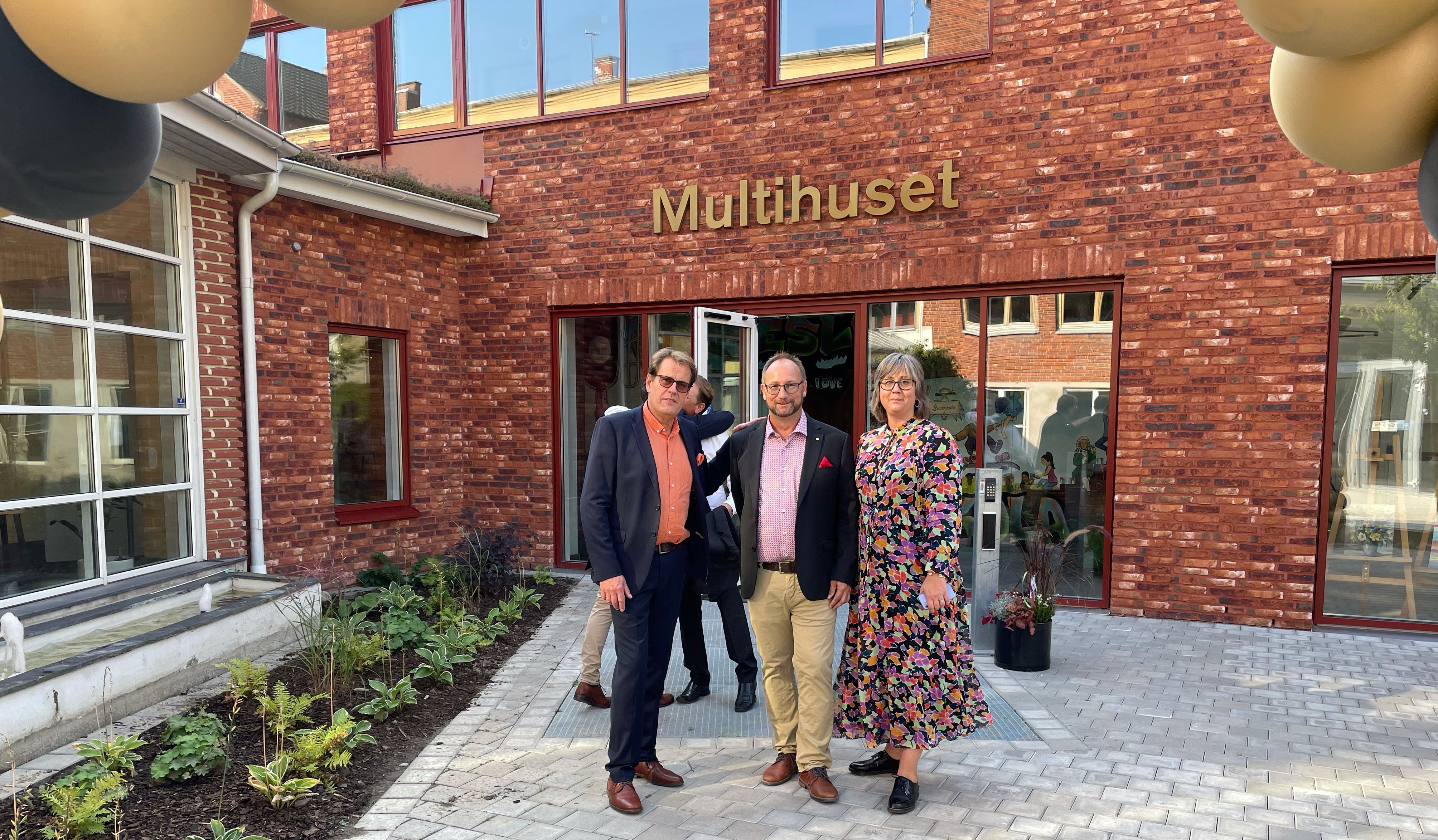 Tv&aring; m&auml;n och en kvinna framf&ouml;r Multihuset i r&ouml;tt tegel. Plan.tering till v&auml;nster