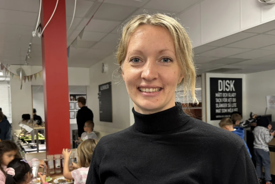Blond kvinna i svart tröja i skolmatsal. I bakgunden syns skolmatsalen och några barn.