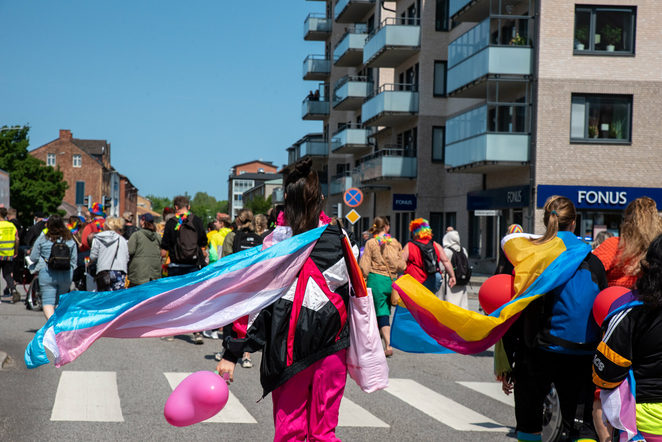 En deltagare i pridetåget syns bakifrån, den håller i en rosaballong och en transgender-flagga