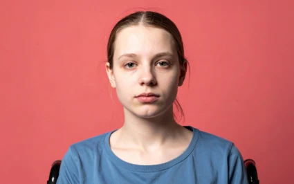 En person sitter framför en korallröd bakgrund. Personen bär en blå t-shirt och delar av en rullstol syns bakom axlarna.