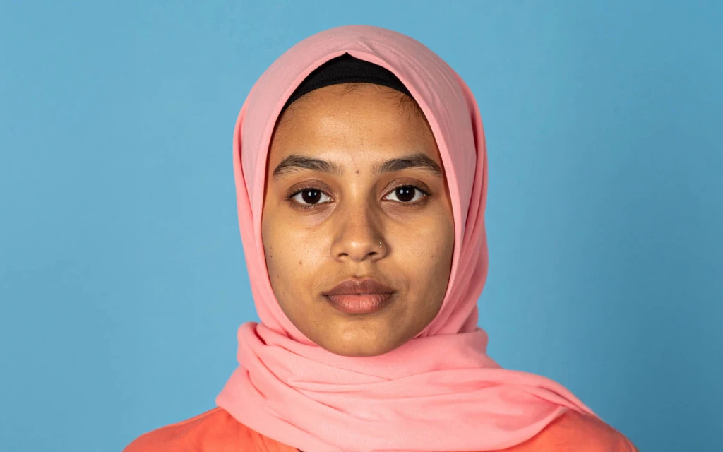 En person står framför en ljusblå bakgrund. Personen bär en rosa hijab och en rosa topp.