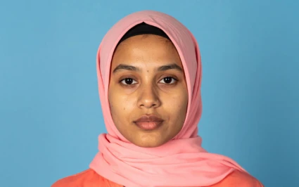 En person står framför en ljusblå bakgrund. Personen bär en rosa hijab och en rosa topp.
