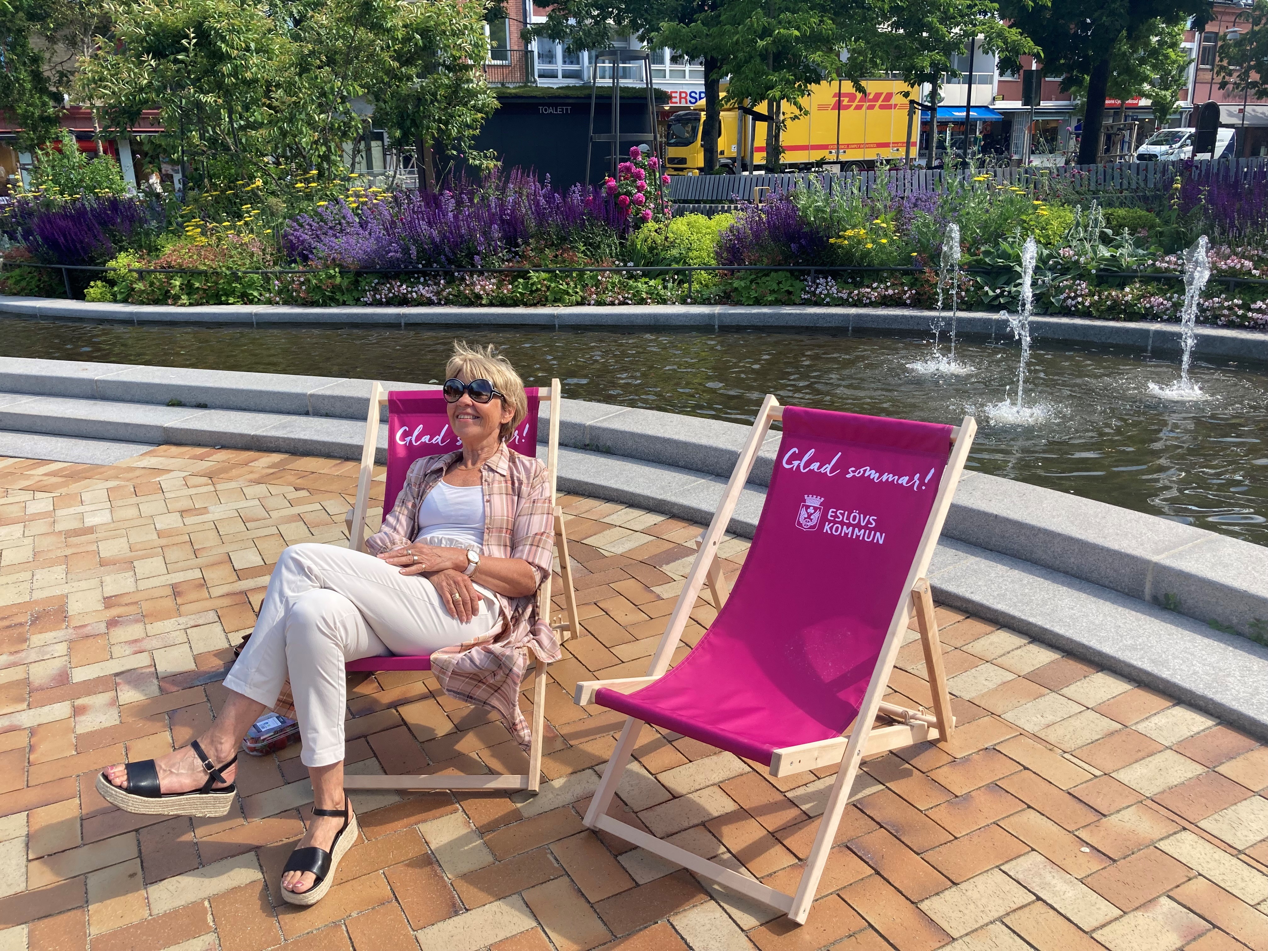 Två rosa solstolar framför dammaen. Frodig växklighet bakom dammen. I en av solarna sitter en blond kvinna i ljusa kläder och med solglasögon.