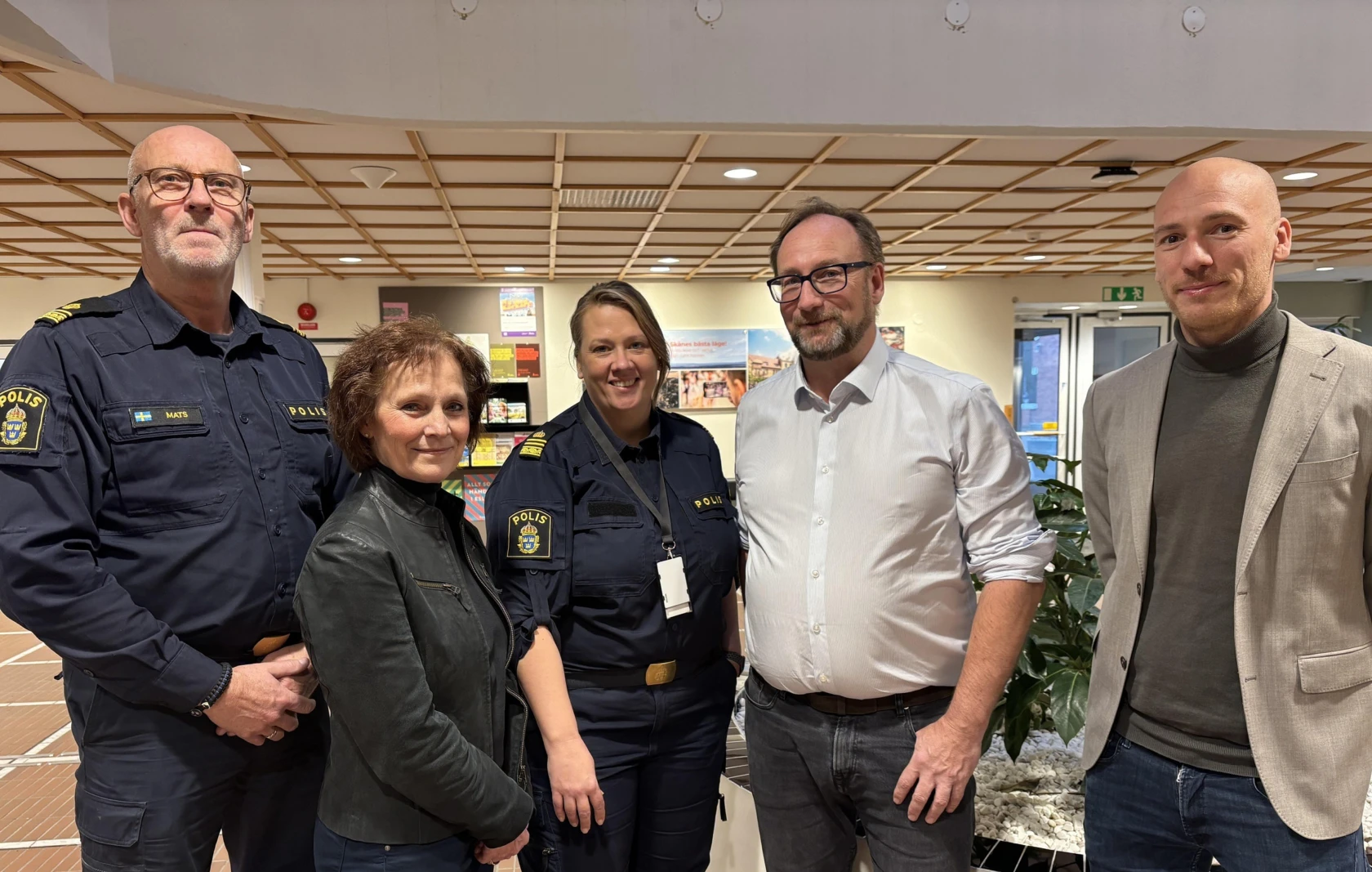 Fem personer, två kvinnor och tre män, står i ett reception och tittar in i kameran. Två av dem bär polisuniform.