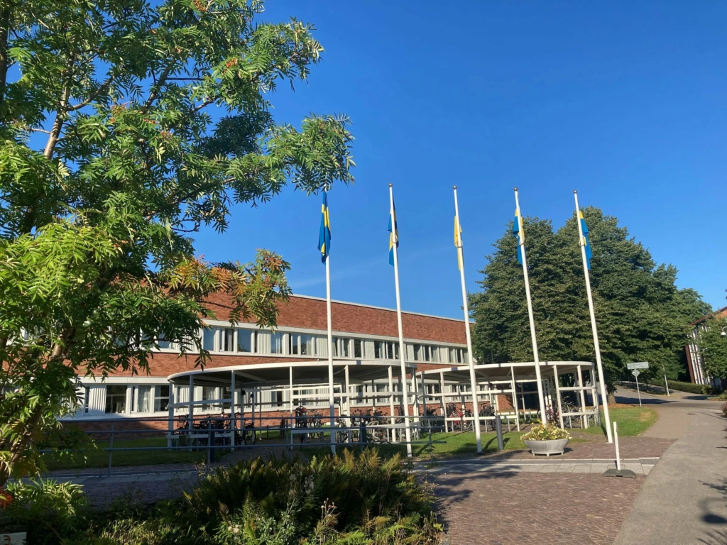 Ukarinas flagga i mitten av fyra svenska flaggor´framför stadshuset. Blå fimmel och gröna träd i förgrunden och bakgrunden.