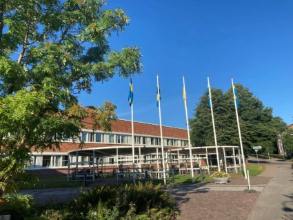 Ukarinas flagga i mitten av fyra svenska flaggor´framför stadshuset. Blå fimmel och gröna träd i förgrunden och bakgrunden.