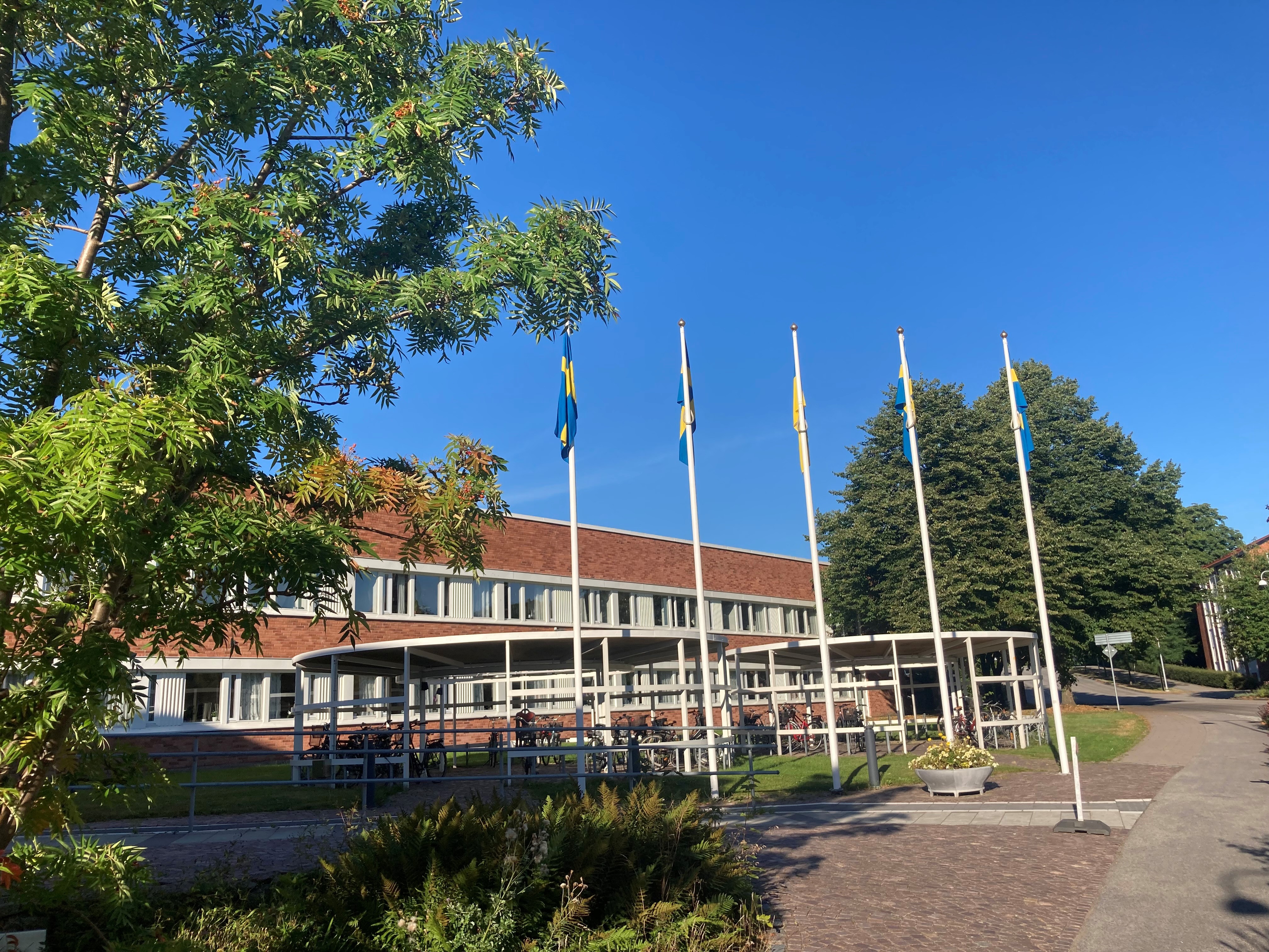 Ukarinas flagga i mitten av fyra svenska flaggor&acute;framf&ouml;r stadshuset. Bl&aring; fimmel och gr&ouml;na tr&auml;d i f&ouml;rgrunden och bakgrunden.