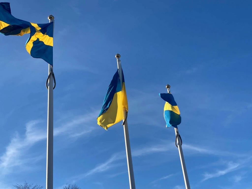Tre flaggstänger mot blå himmel. Sveriges flagga på två av dem, Ukrainas flagga i mitten.