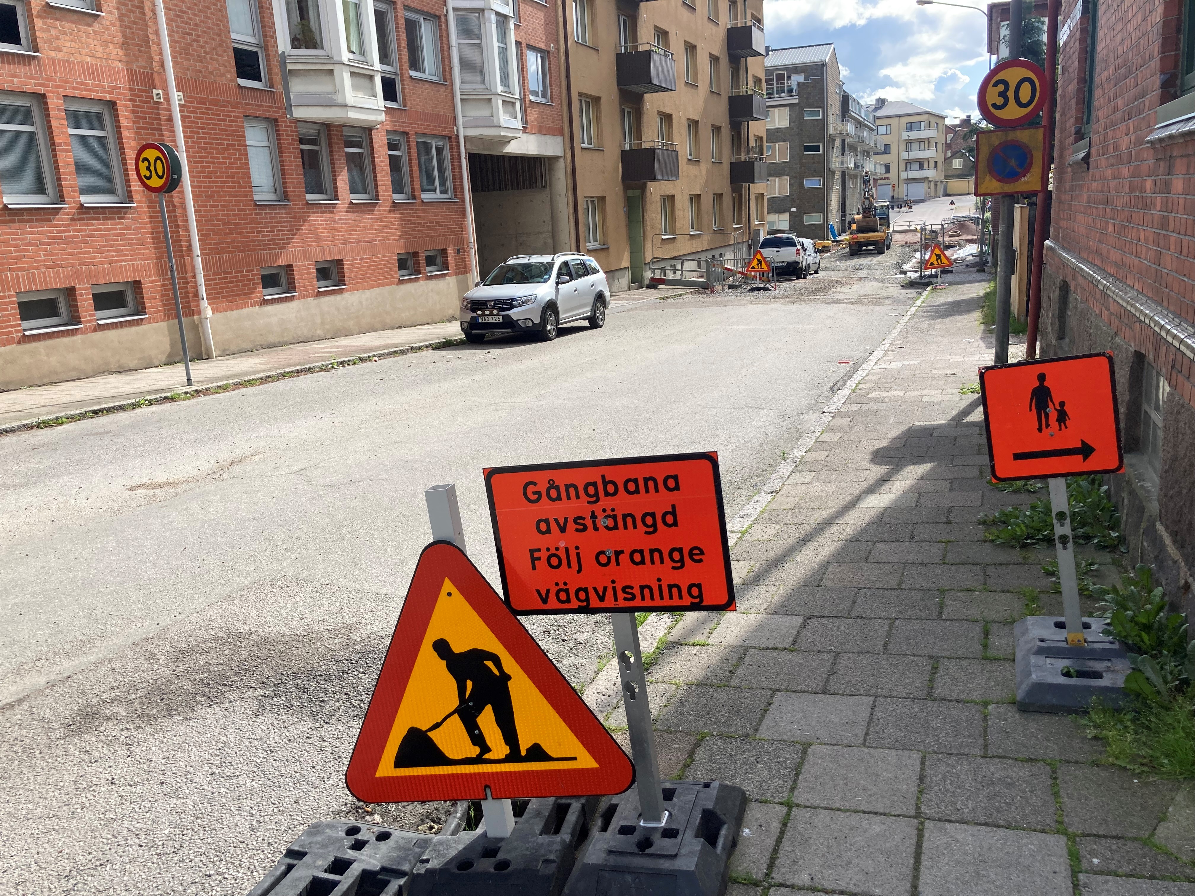 Varningstriangel och r&ouml;d skylt med text att gatan &auml;r avst&auml;ngd. Gata med parkerade bilar och tegelhus bakom.